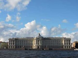 A grand classical building by the water under a blue sky with clouds.