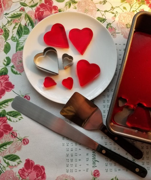 Heart-shaped cookie cutters and red heart cutouts on a plate with a knife and spatula nearby.
