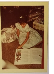 Young girl in a blue dress kneeling on the floor reading a book.