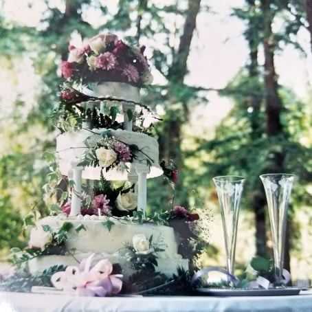 Elegant three-tier wedding cake decorated with flowers on a table outdoors.