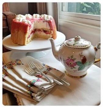 A layered cake with pink frosting and cream filling beside a floral teapot.