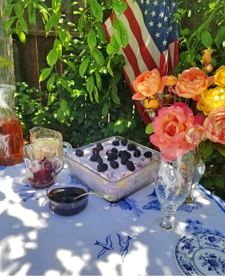 A dessert table with a blueberry cake, flowers, and an American flag outdoors.