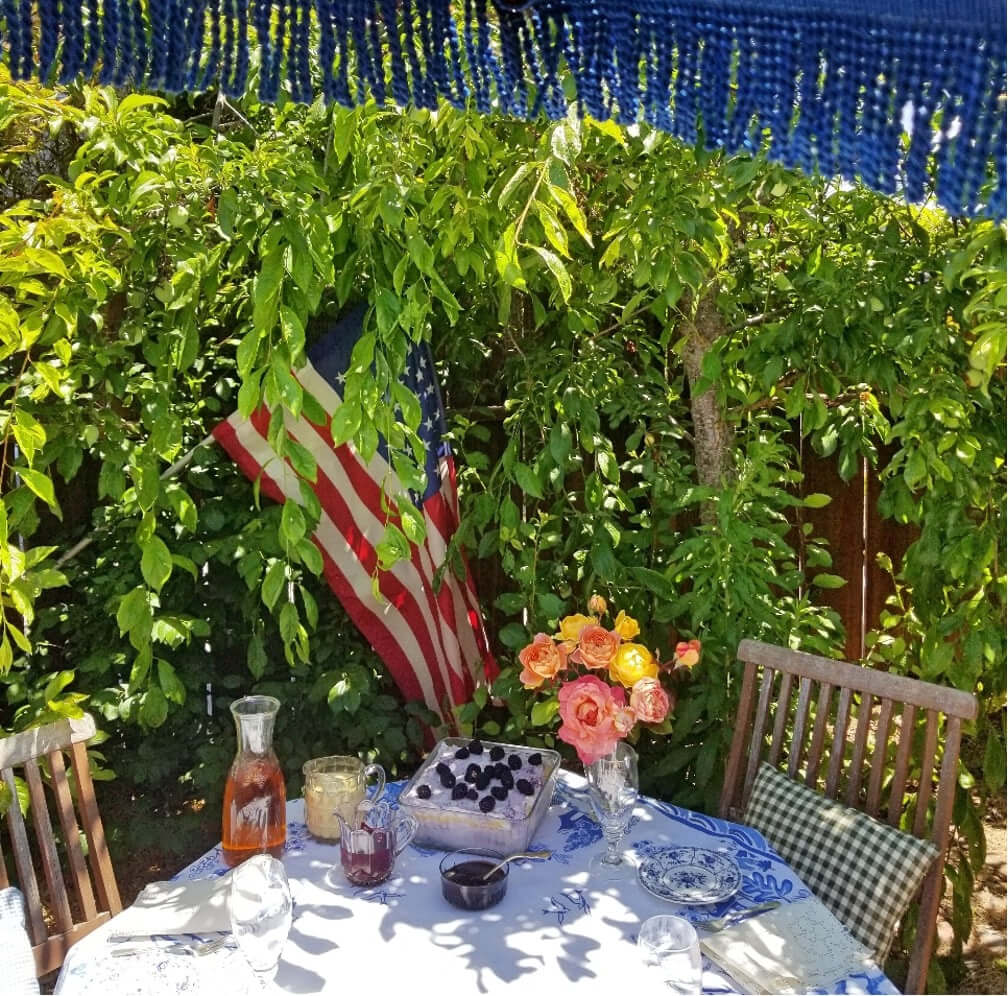 A shaded outdoor table with flowers, drinks, and an American flag in the background.
