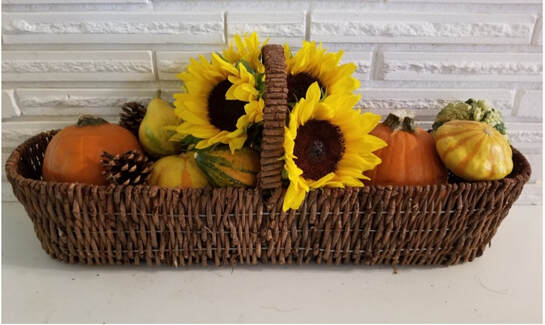 Sunflowers and small pumpkins in a woven basket against a white brick background.