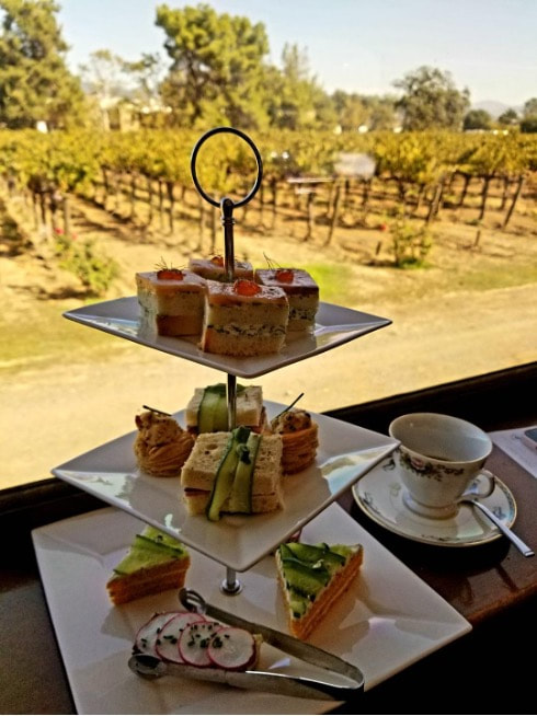 Three-tiered afternoon tea set with sandwiches, pastries, and tea in a vineyard setting.