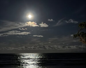 Full moon shining over a calm ocean with clouds scattered in the night sky.