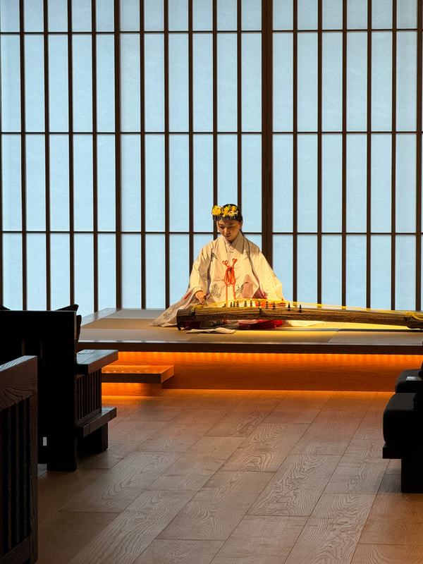 A person in traditional attire sits cross-legged on a wooden platform in a minimalist room.