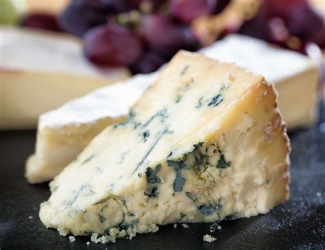 A wedge of blue cheese with visible blue veins and grapes in the background.