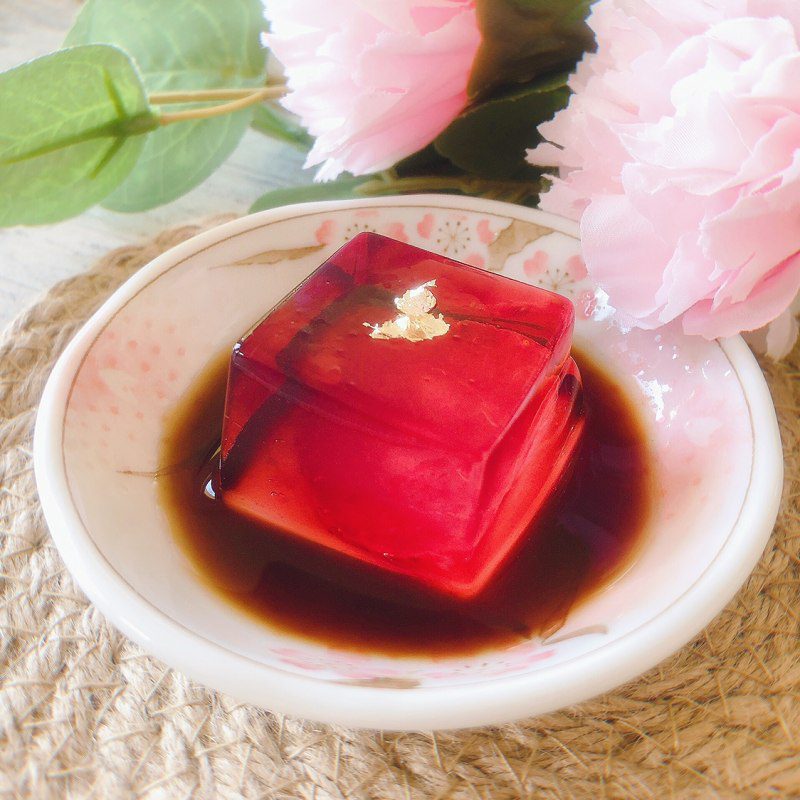 A translucent red cube dessert with gold leaf on top, served in a small dish with sauce.