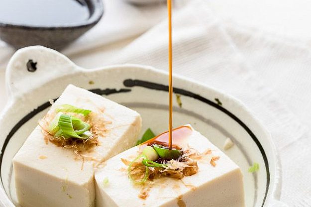 Soy sauce being poured over tofu garnished with green onions and bonito flakes.