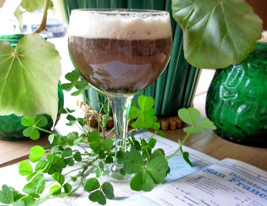 A glass of layered coffee drink with frothy top, surrounded by green plants.