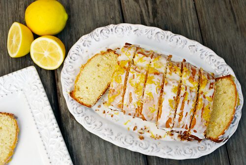 Lemon drizzle cake sliced on a white plate with lemons beside it.