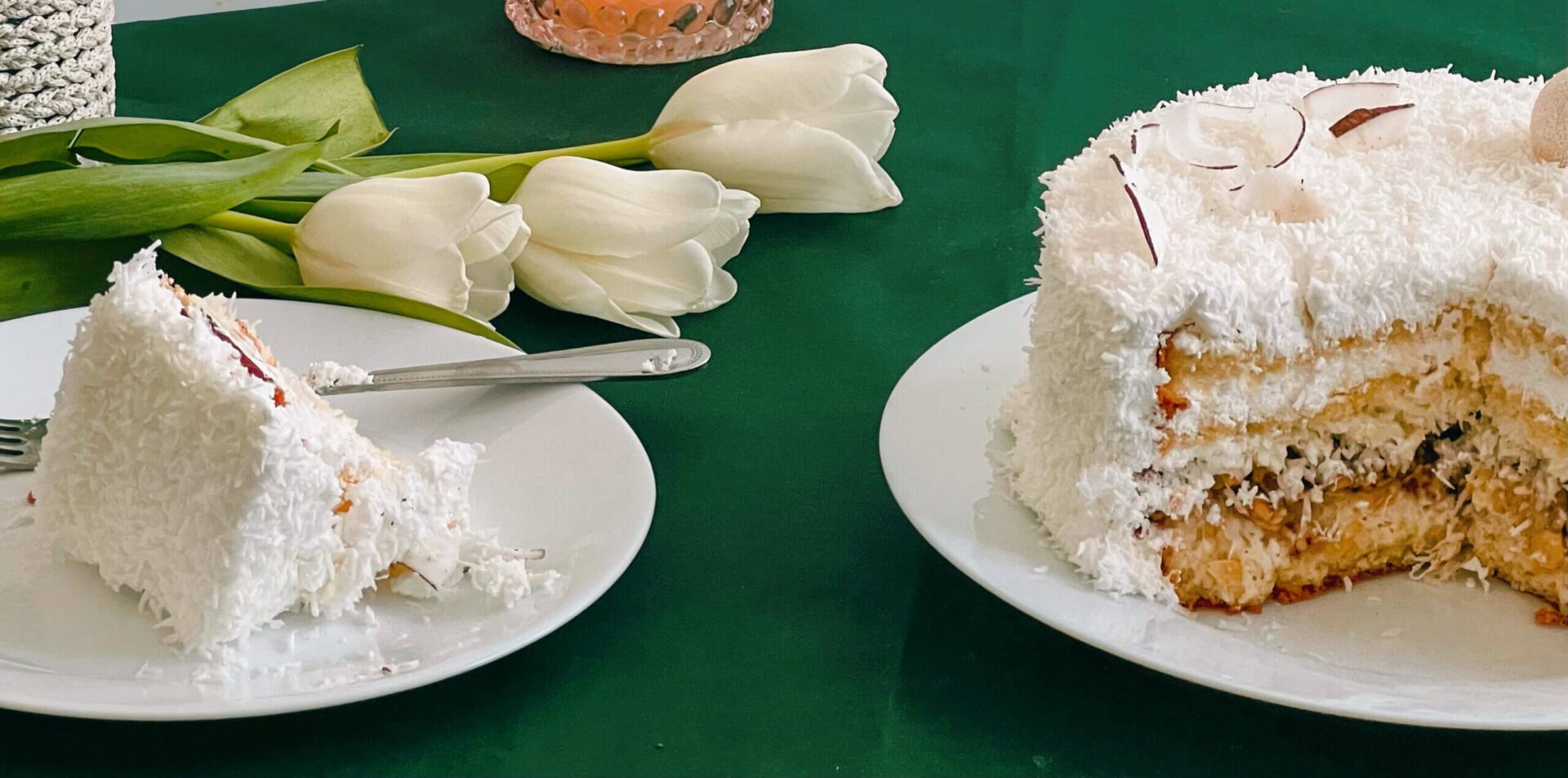 A slice of coconut cake on a plate beside the remaining cake on a green table.