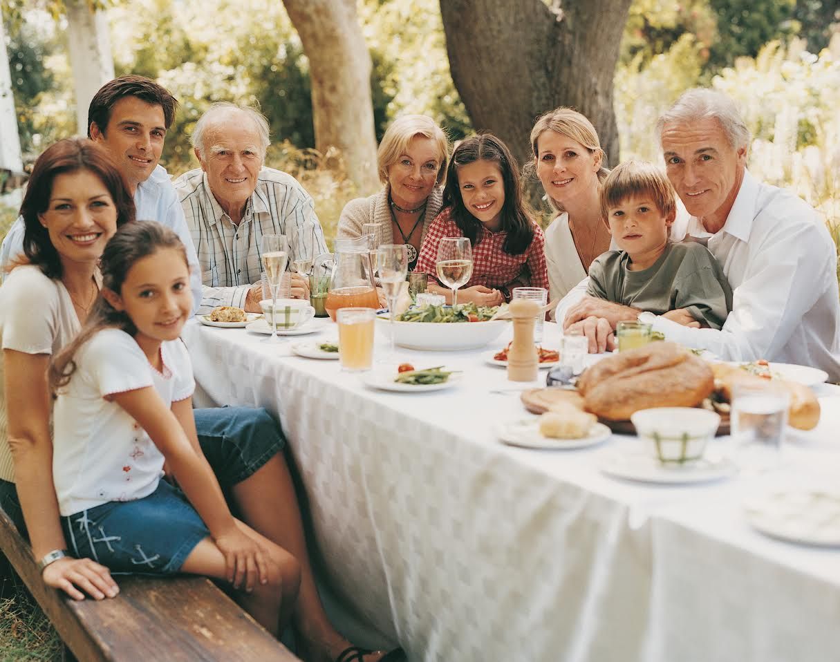 Family enjoying a meal together outdoors at a long table under trees.