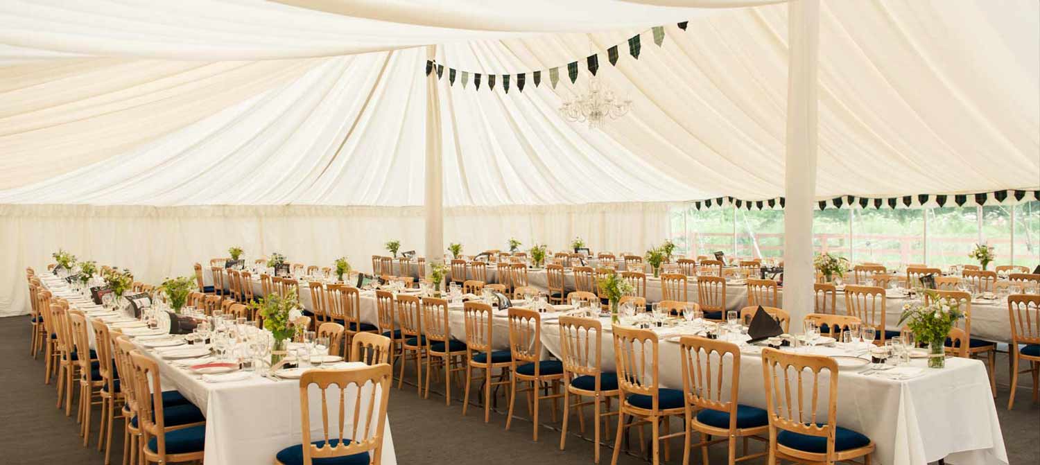 Elegant wedding reception setup inside a white tent with wooden chairs and floral centerpieces.