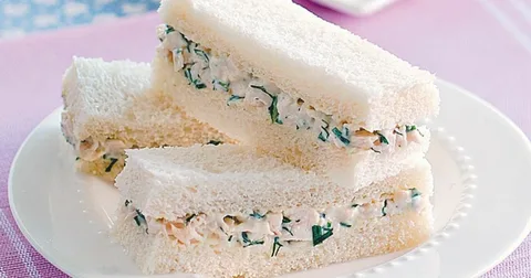 Close-up of a creamy chicken salad sandwich with herbs on white bread.