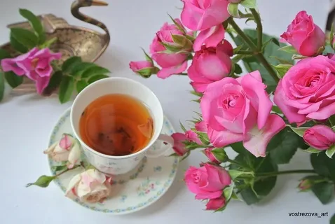 A cup of tea beside vibrant pink roses on a white surface.