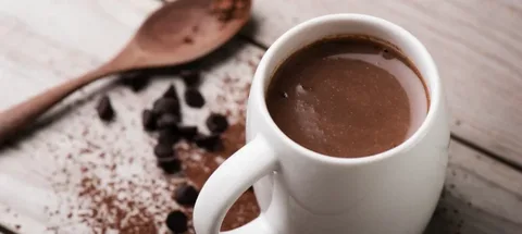 Close-up of a white cup filled with hot chocolate on a dark surface with cocoa beans.