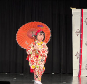 A young child in traditional Japanese attire holding a red parasol on stage.