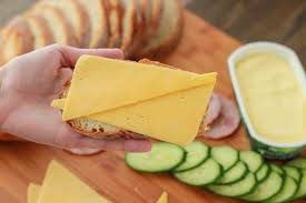 A hand holding a sandwich with cheese and ham, cucumber slices on the side.