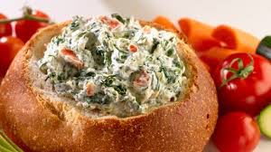 Creamy spinach dip served in a bread bowl with carrot sticks.