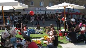 People relaxing on grass in a busy urban area with umbrellas and buildings.