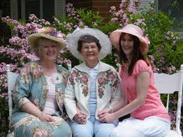 Three women in floral hats and pastel outfits smiling outdoors.