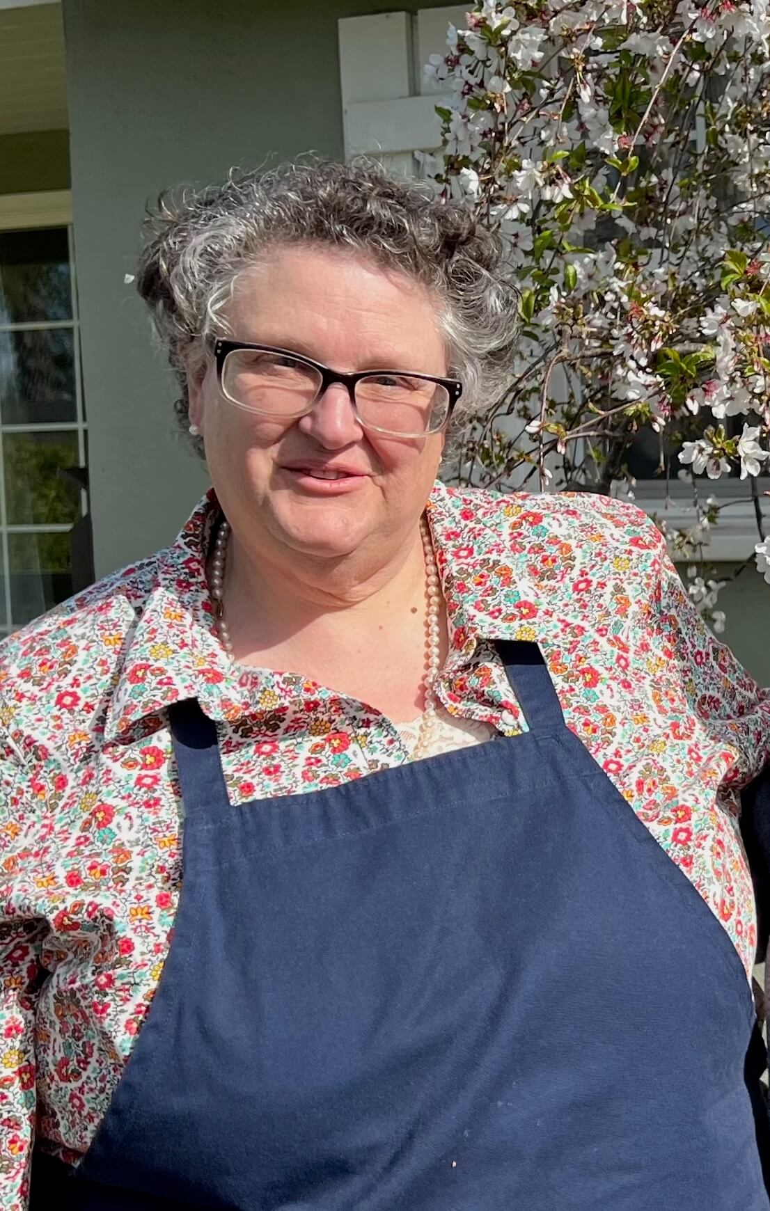 A smiling woman wearing glasses and a floral shirt with a navy apron.