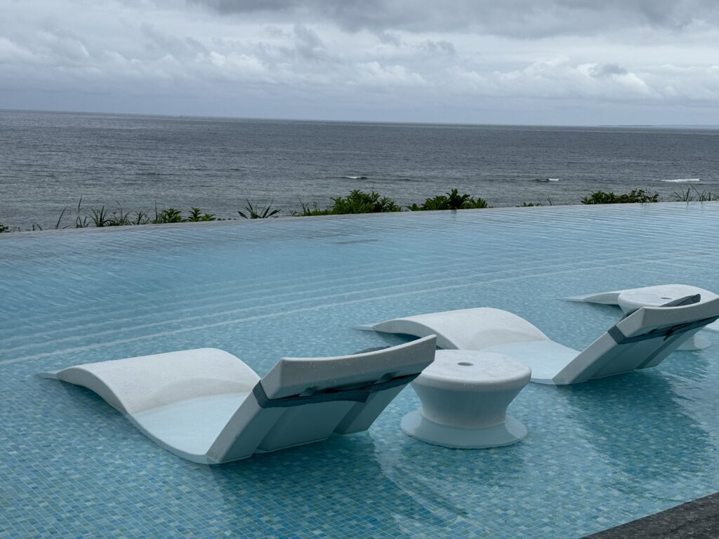 Modern white chairs and table in a pool with ocean view under cloudy sky.