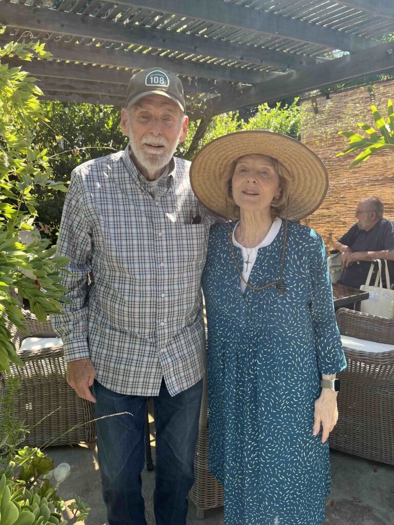 Elderly couple smiling outdoors in casual attire and sun hat.